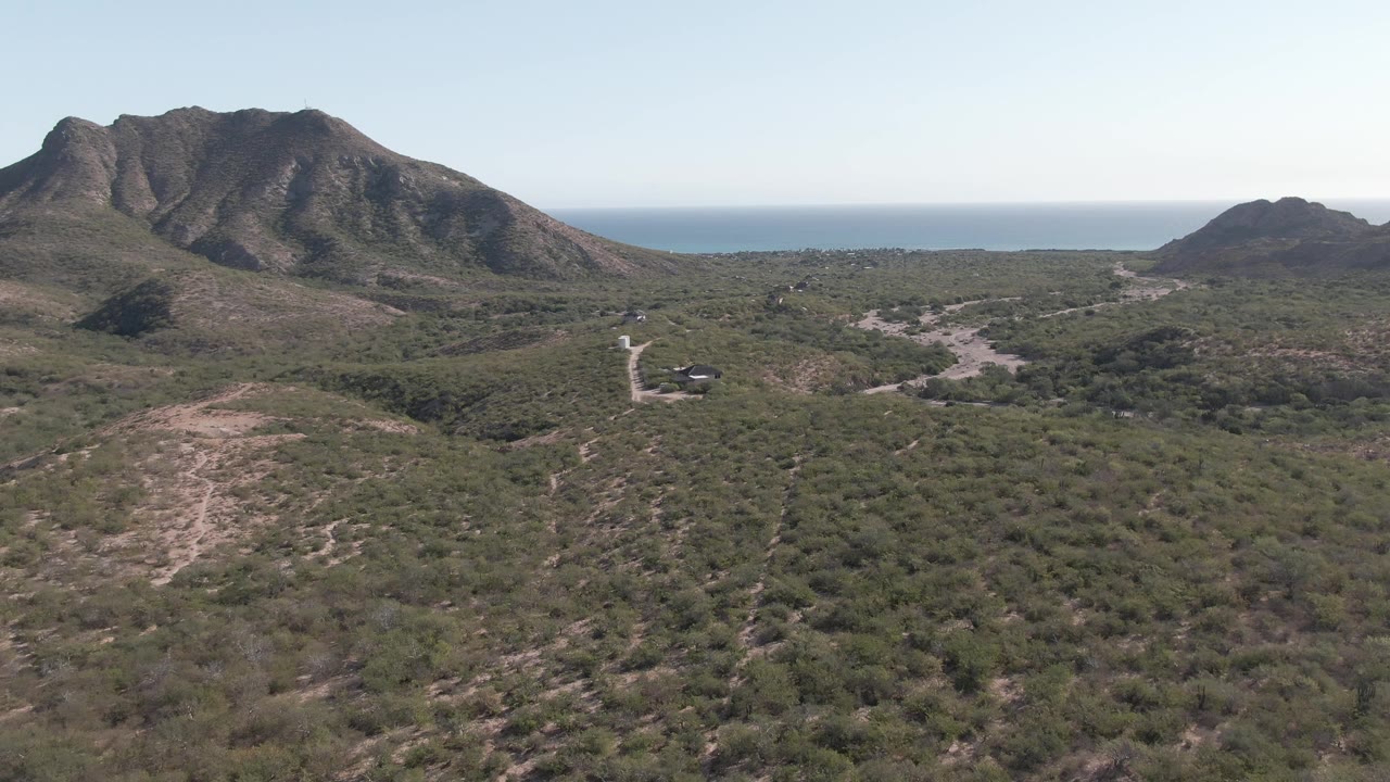 Aerial view in Cabo Pulmo, Baja California Sur, with dry scrubland in the foreground, a winding hillside road, rugged mountains, a thin ocean horizon, and no visible people.