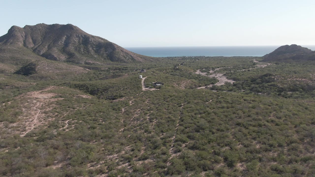 One second into the Cabo Pulmo clip, the elevated frame shows the pale road crossing scrubby hills toward dark mountain ridges, with the ocean line behind them and no visible people.