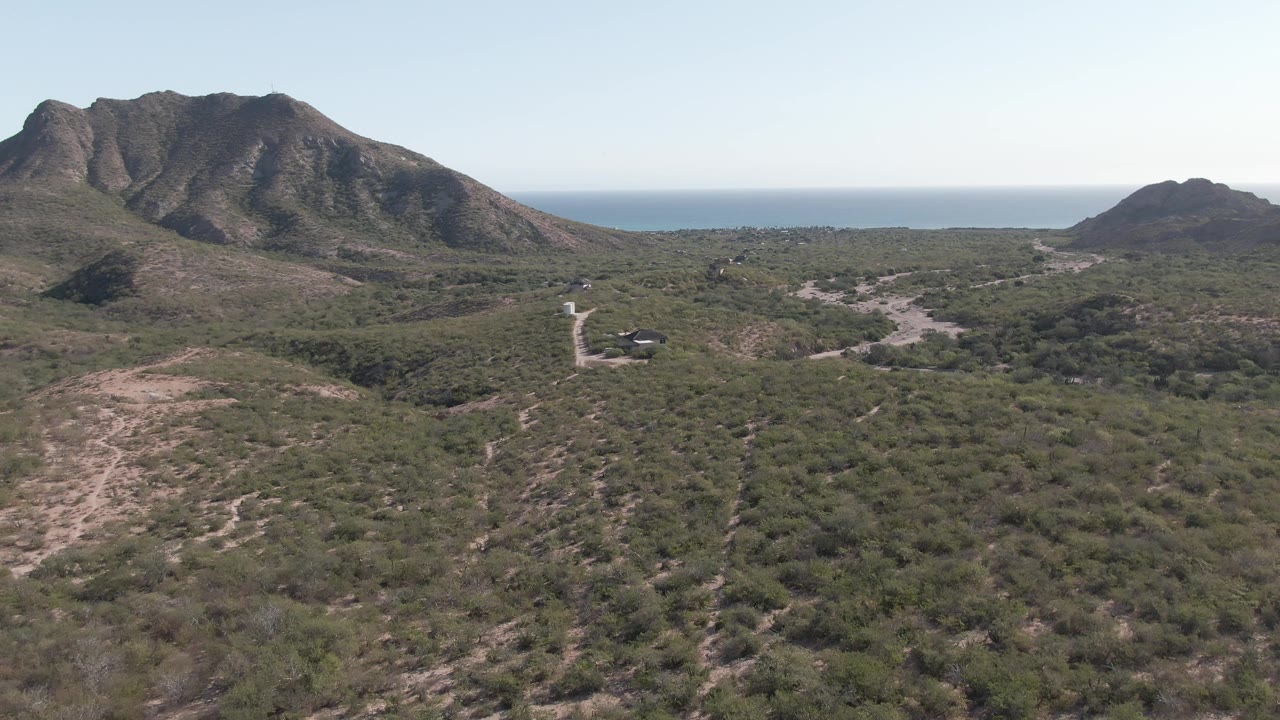 The Cabo Pulmo aerial view continues with a broad foreground of dry vegetation, the road and vehicle near the center-right, mountains across the horizon, ocean beyond, and no visible people.