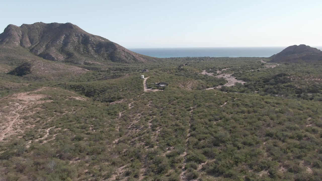 At three seconds, the aerial composition places the winding road and small vehicle low in the hillside terrain, with green-brown brush, rugged mountains, distant ocean, and no visible people.