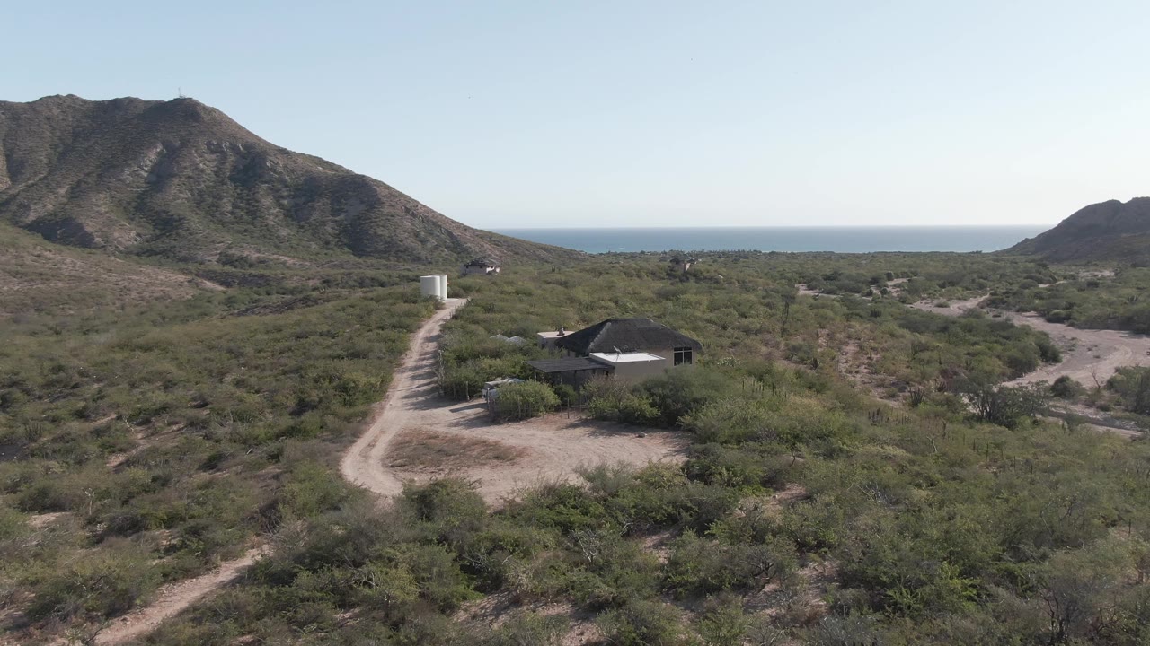 Aerial frame from Cabo Pulmo shows a dirt road leading past a small house through desert scrub toward low mountains and a distant ocean horizon, with no visible people.