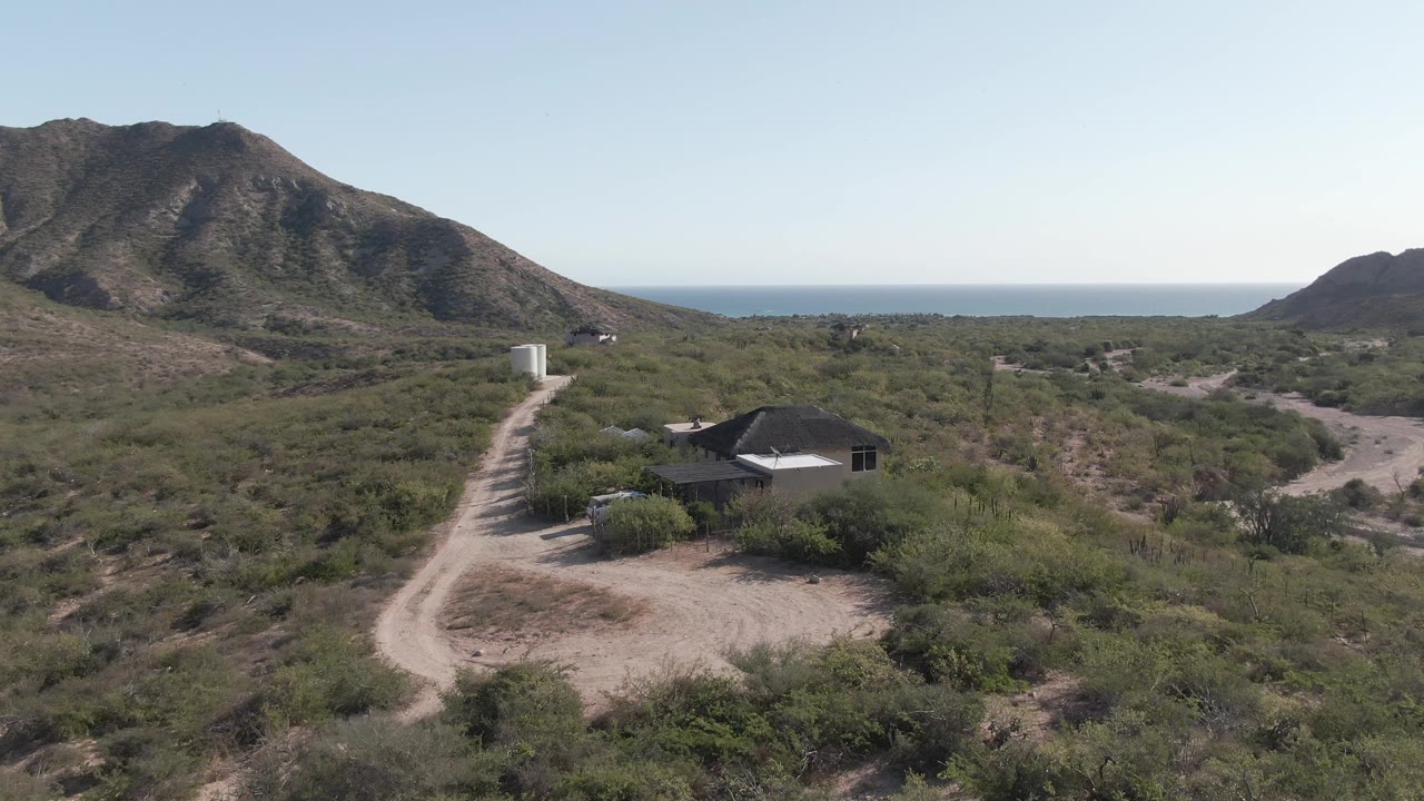 Cabo Pulmo aerial view shows the dirt road and white tank more centered as the small house sits among green desert vegetation below rugged hills, with no visible people.