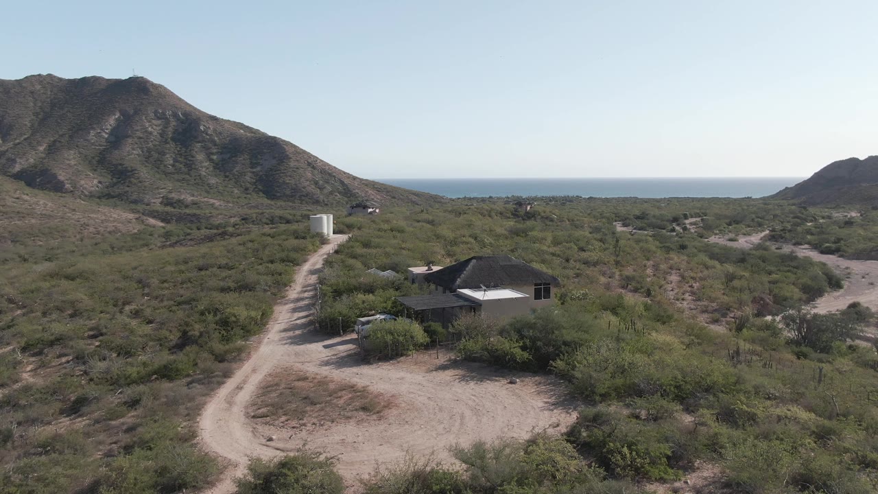 The third Cabo Pulmo frame holds a wide elevated composition with the road crossing the scrubland toward the house and a blue ocean line beyond the mountains, with no visible people.