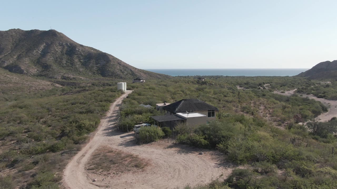 Closer aerial view of a dirt driveway, house, parked vehicle, desert scrub, and mountain backdrop.