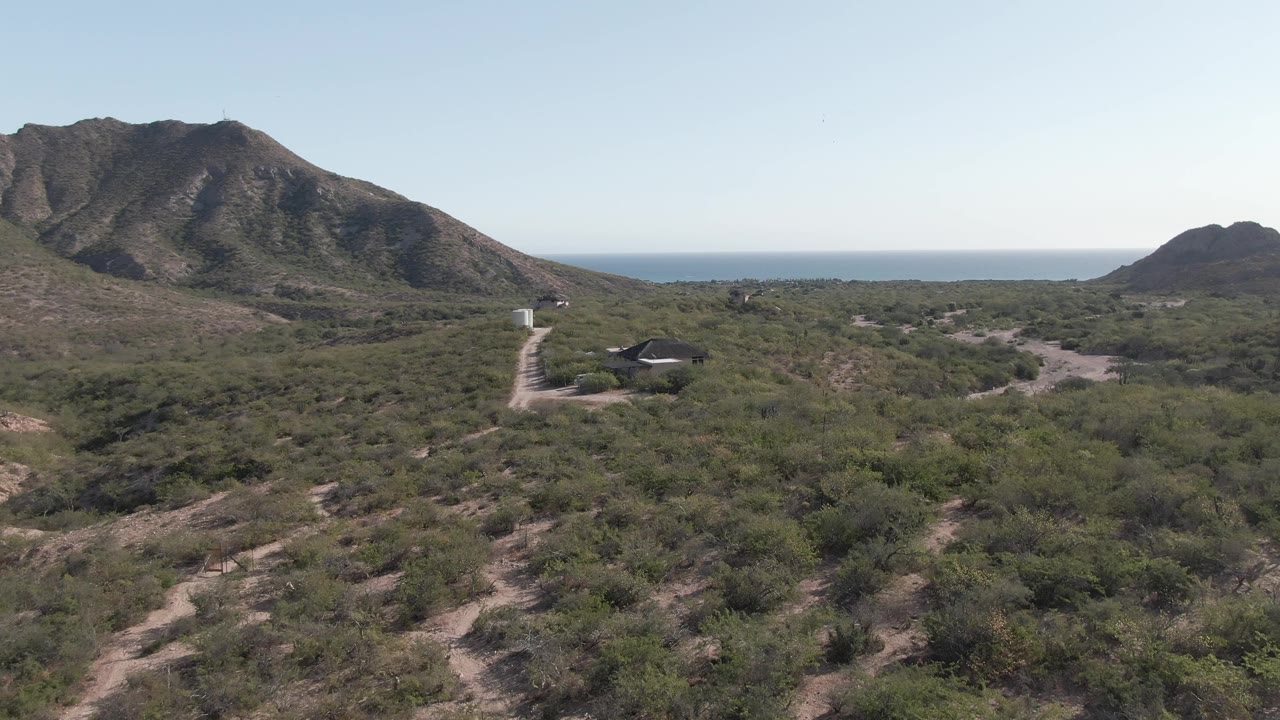 Aerial frame in Cabo Pulmo showing sandy scrubland, a pale dirt road curving past a dark-roofed structure, mountains, distant ocean, and no visible people.