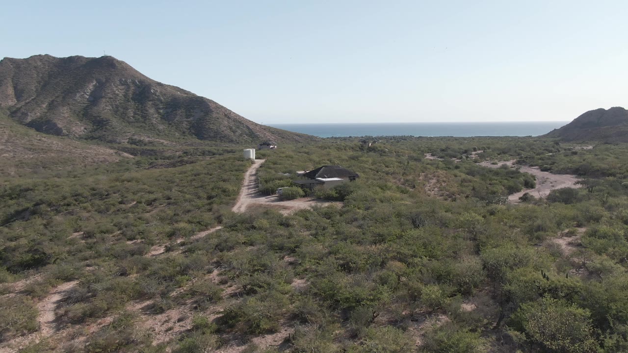 Later aerial frame over Cabo Pulmo with dry green scrub in the foreground, a winding dirt road at left, a dark-roofed structure in the midground, mountains, ocean, and no visible people.