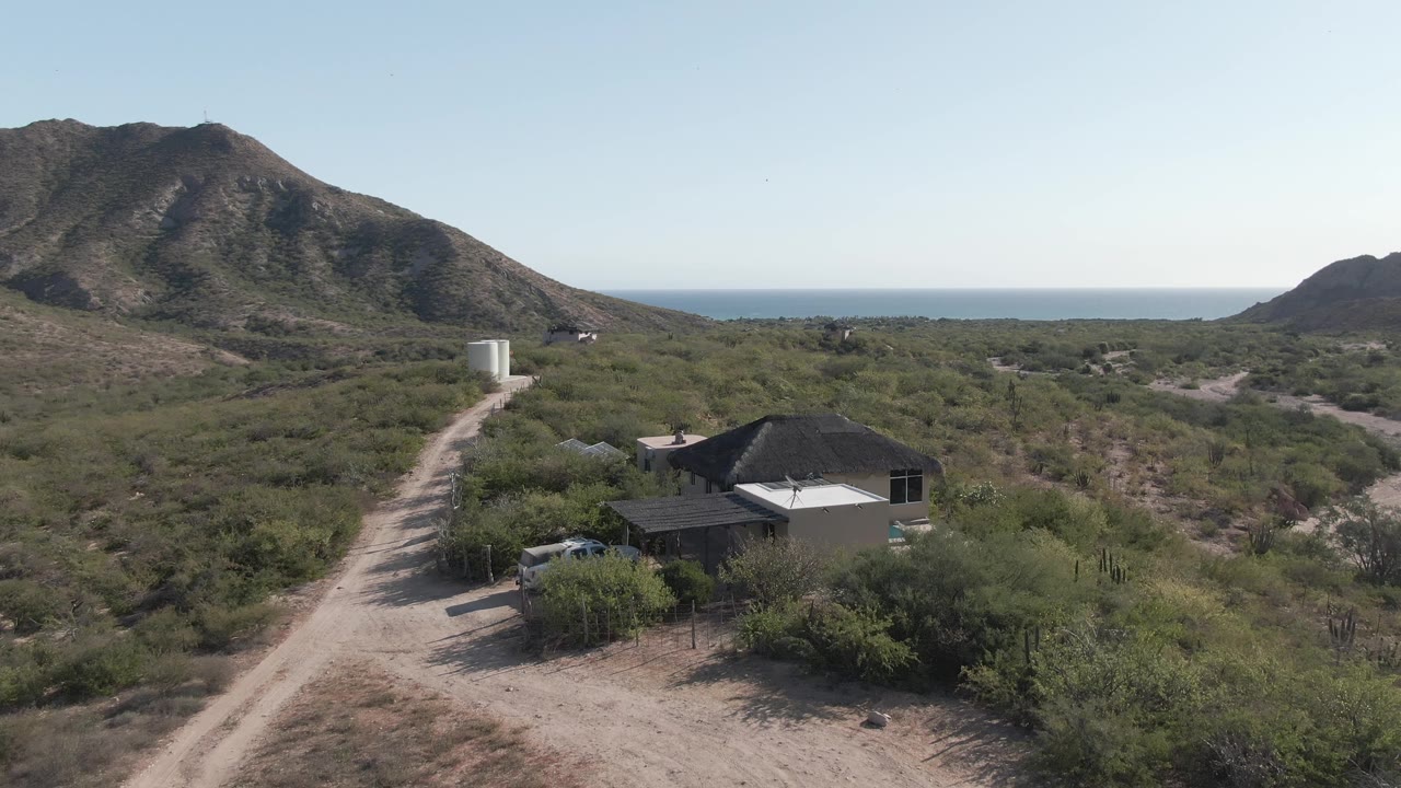 Opening frame over Cabo Pulmo, Baja California Sur, showing a low desert house beside a pale dirt road, scrub vegetation, a mountain backdrop, ocean horizon, and no visible people.