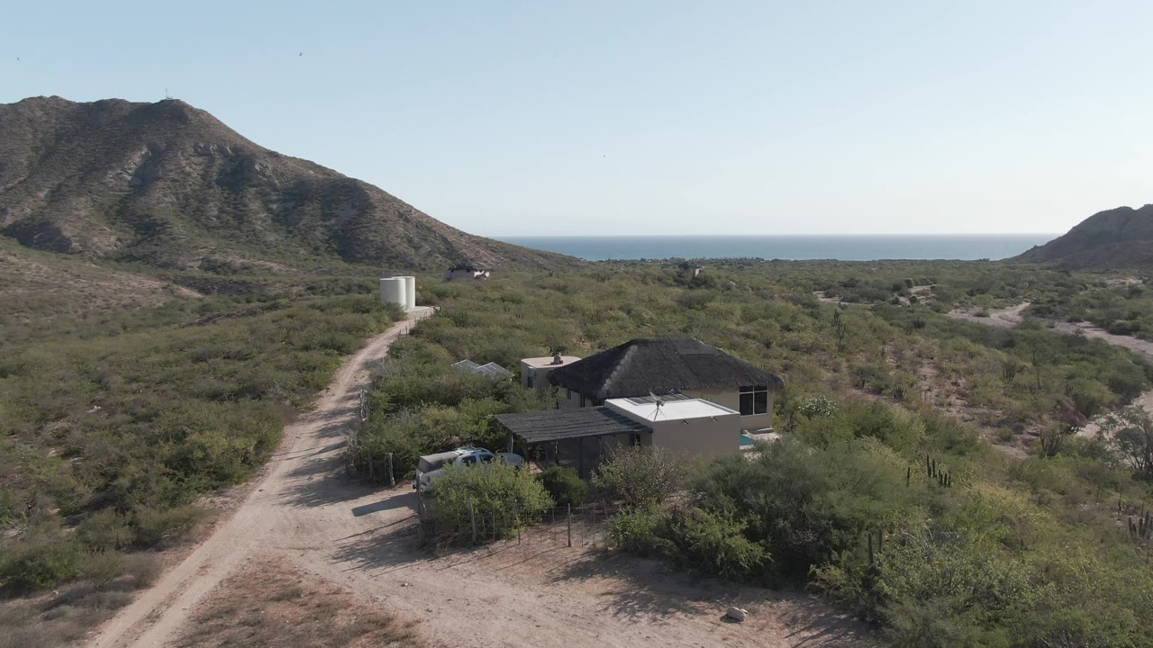 At one second, the Cabo Pulmo aerial view keeps the desert house near the lower center-right, with a dirt road, brush-covered terrain, mountain ridge, ocean line, and no visible people.