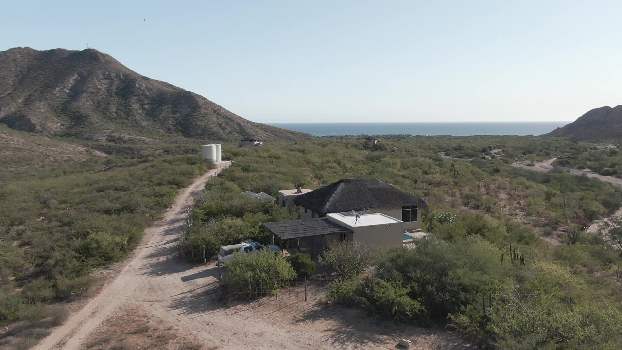 At two seconds, the Cabo Pulmo frame shows the house slightly farther along the drone view, a white vehicle partly hidden by brush, a winding dirt road, the mountain, ocean horizon, and no visible people.