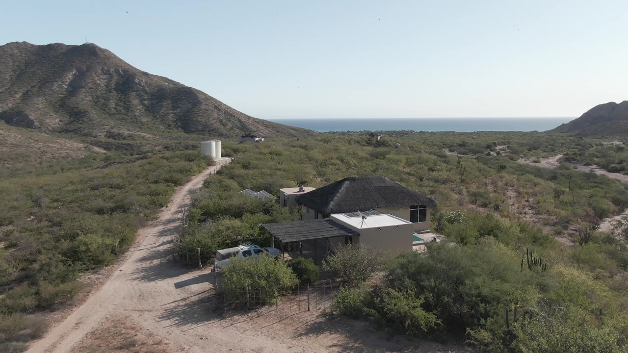 Aerial frame of desert brush, a dirt road, a house, mountains, and ocean horizon.