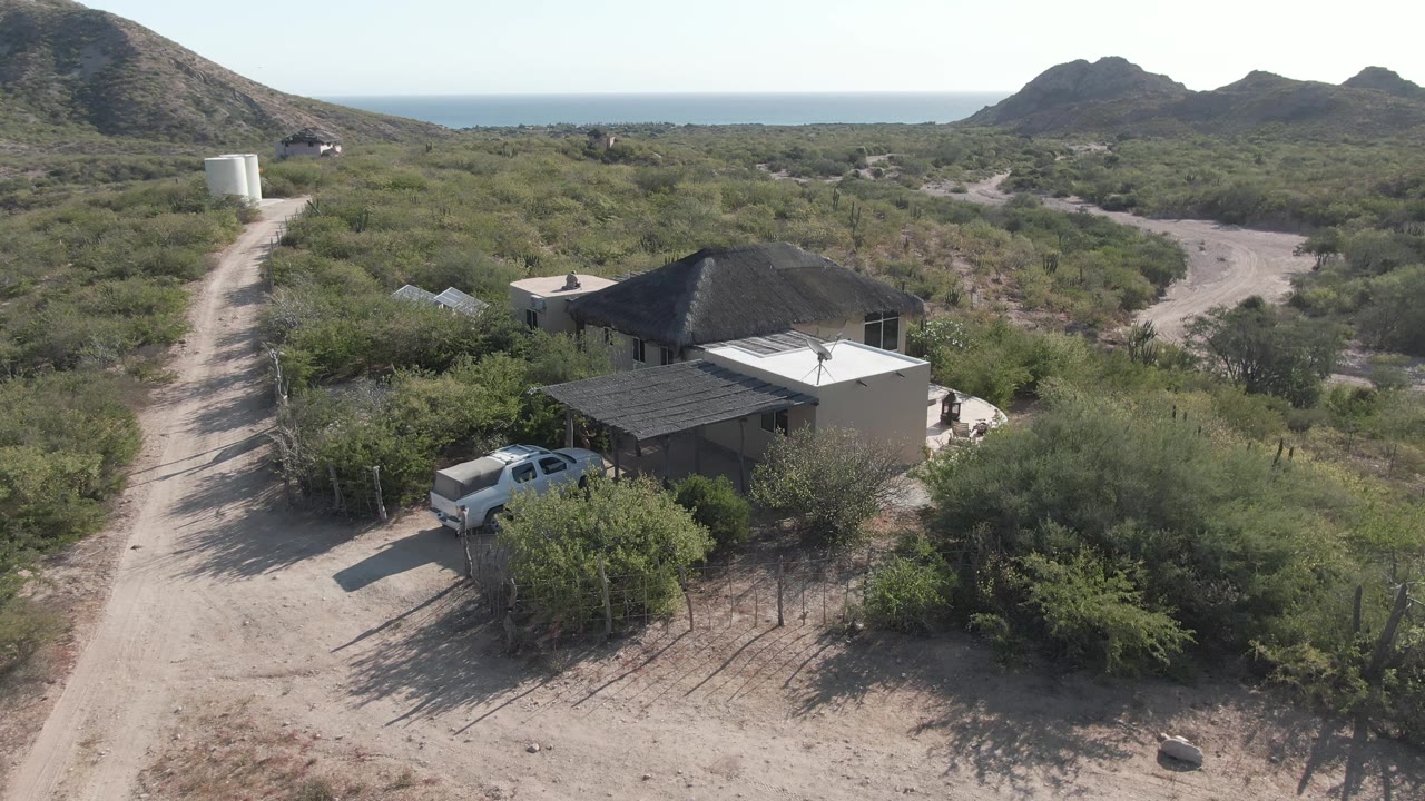 Opening aerial frame over Cabo Pulmo shows a small desert house near a white truck, a pale dirt road, scrub vegetation, low hills, and a distant ocean horizon with no visible people.