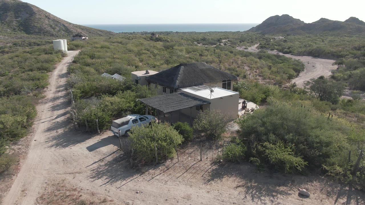 Drone view of a white truck by a rural desert house and dirt road near hills.