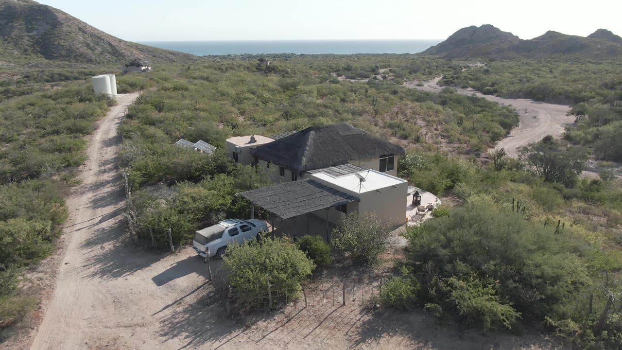 At 0:02, the drone angle shows the white truck near the house and shaded structure, with the dirt road leading past a white tank into scrubland, hills, and the ocean line in Cabo Pulmo, with no visible people.