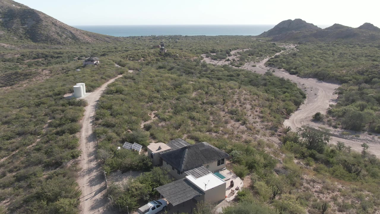 Aerial frame in Cabo Pulmo, Baja California Sur shows a house near the bottom edge, a car by the road, scrub-covered hills, a winding dirt road, distant ocean, and no visible people.