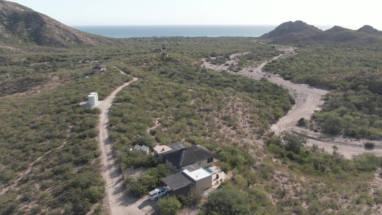 Aerial frame in Cabo Pulmo, Baja California Sur holds a wide view of the house near the lower frame, the car on the adjacent road, branching dirt tracks, scrub vegetation, low mountains, ocean horizon, and no visible people.