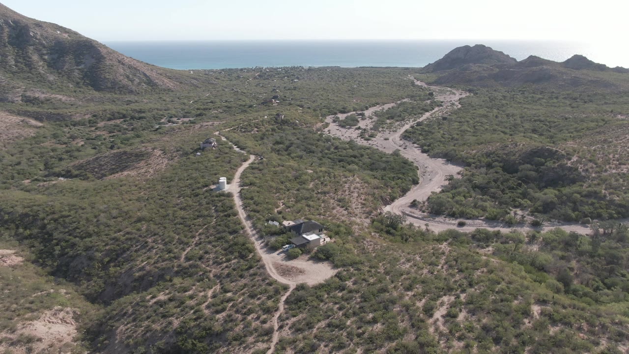 Aerial frame from Cabo Pulmo, Baja California Sur, Mexico showing a winding dirt road, green scrub hills, small structures near the lower left, distant rocky hills, ocean horizon, and no visible people.