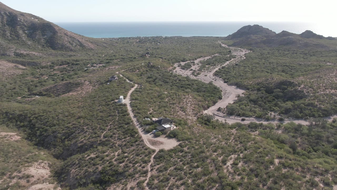 Drone-height frame from Cabo Pulmo showing the pale road curving from the lower left into the center, a small vehicle on the track, low green vegetation, rocky hills, ocean beyond, and no visible people.