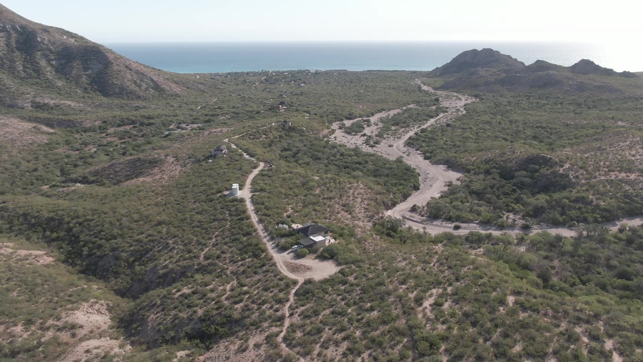 Wide aerial frame in Cabo Pulmo with a dirt road crossing the lower terrain and bending through green scrubland toward dark rocky hills, with the ocean horizon visible and no visible people.