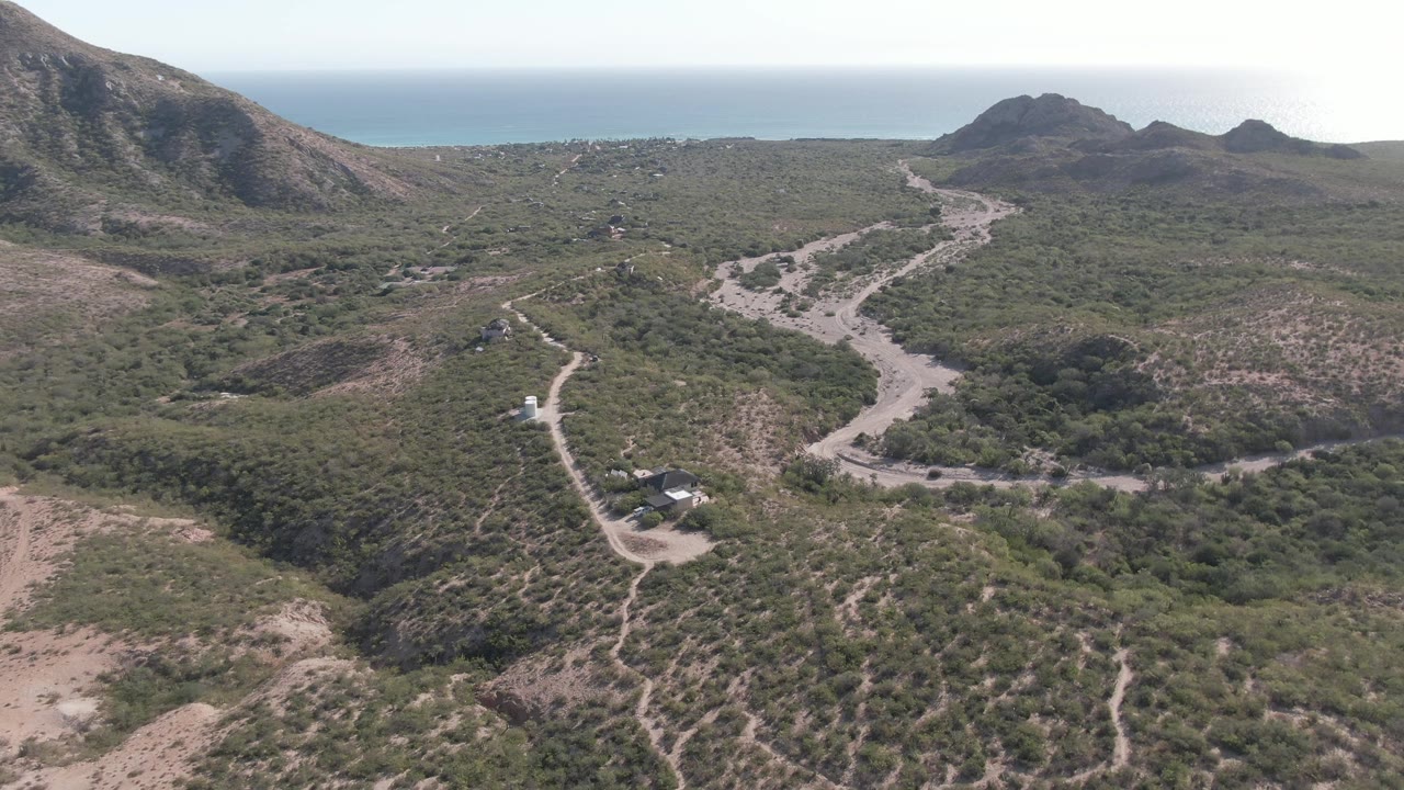 Drone view of a dirt road and small building below rocky hills near the ocean.