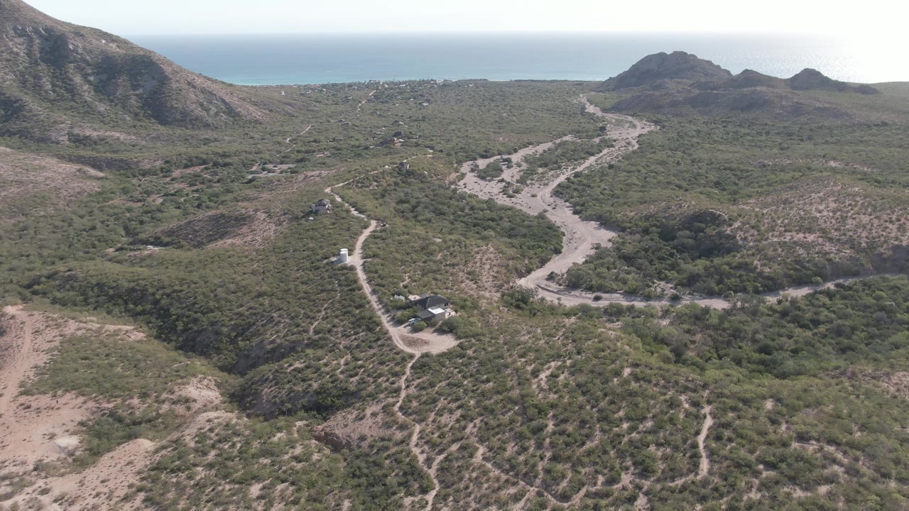 A wide Cabo Pulmo aerial frame shows branching dirt tracks across brushy terrain, a darker tree-filled wash in the midground, mountain ridges, and the ocean line beyond, with no visible people.