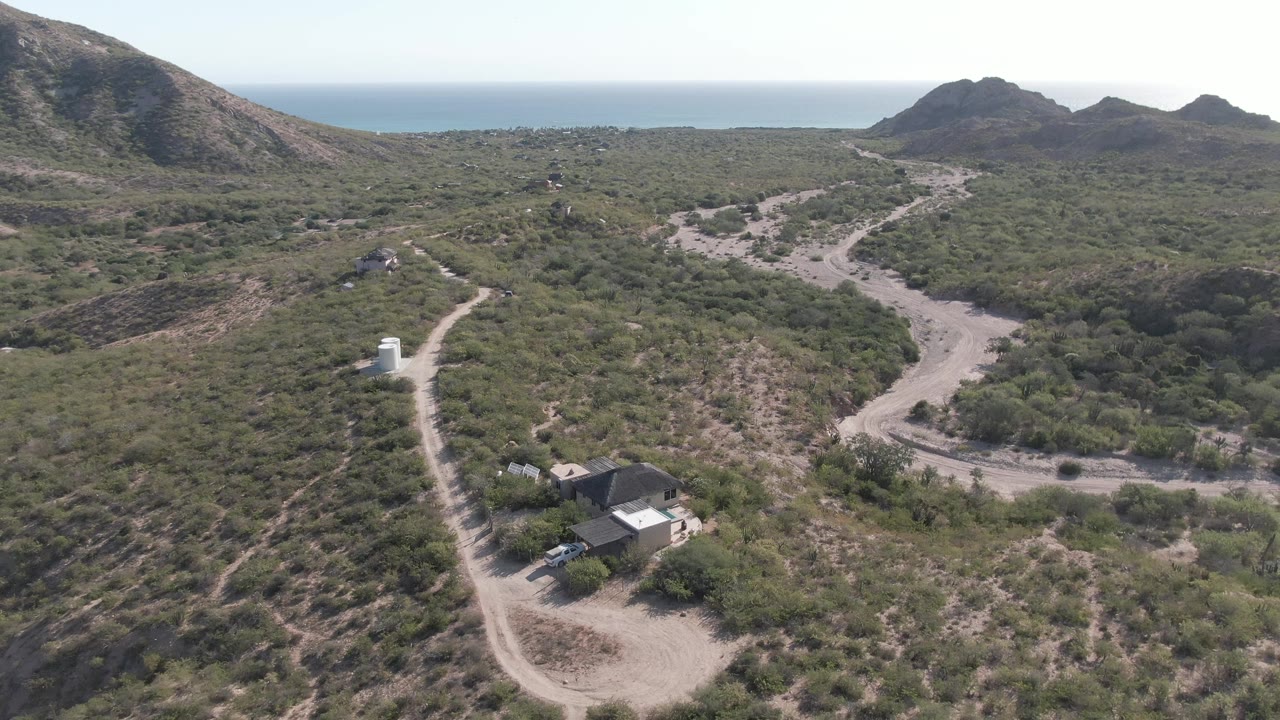 Aerial view over Cabo Pulmo scrubland with winding dirt roads, low hills, a small white roadside structure, distant ocean horizon, and no visible people.