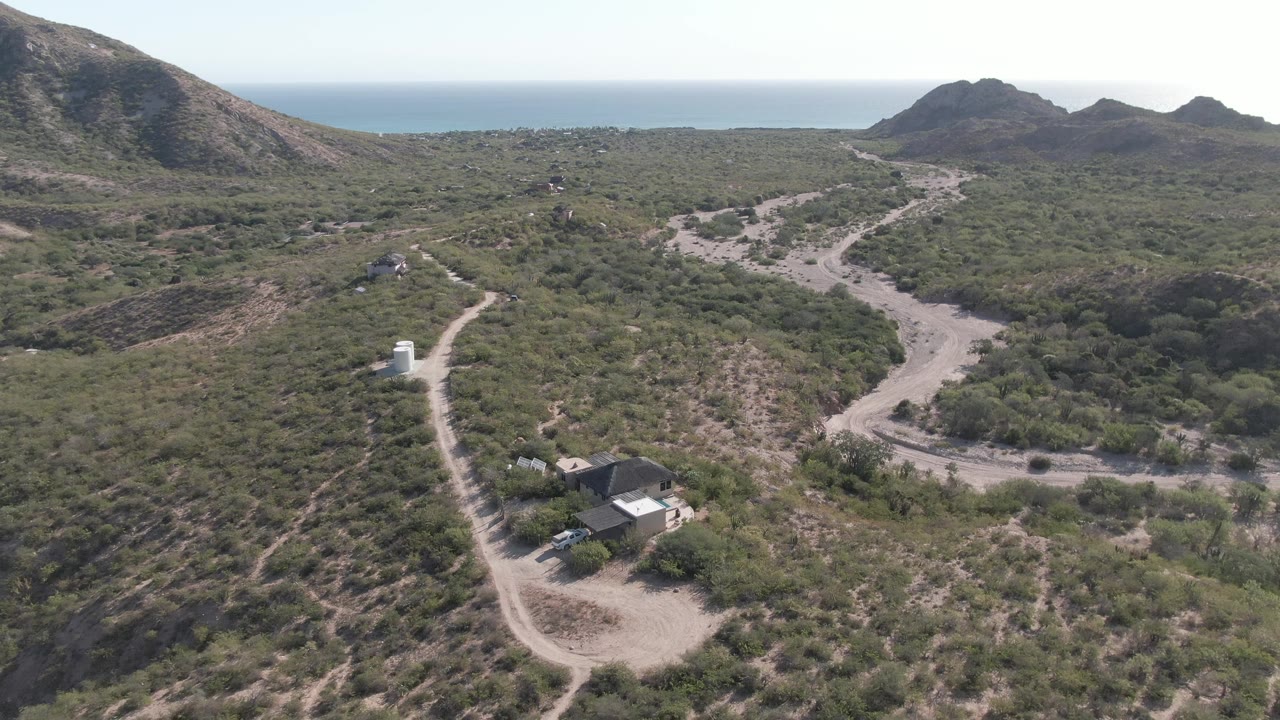 Drone view in Cabo Pulmo with a small building near the lower left, a white roadside structure up the track, branching dirt roads, rugged hills, ocean horizon, and no visible people.