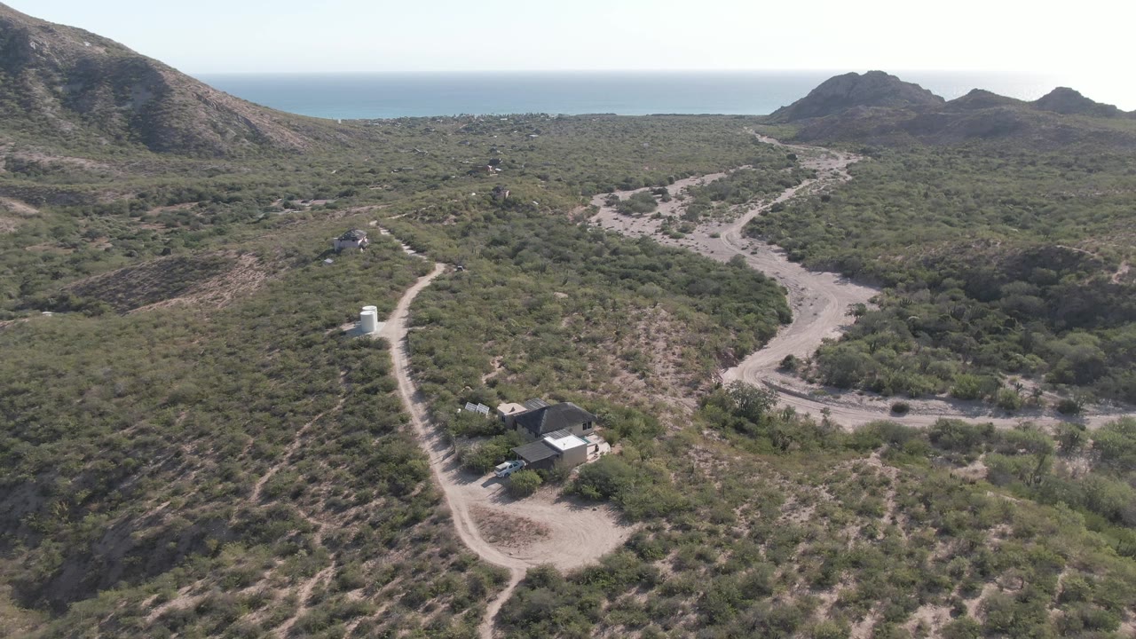 Wide aerial frame of Cabo Pulmo terrain showing tan dirt tracks through green brush, a cluster of small structures near the lower edge, dark hills, blue ocean in the background, and no visible people.