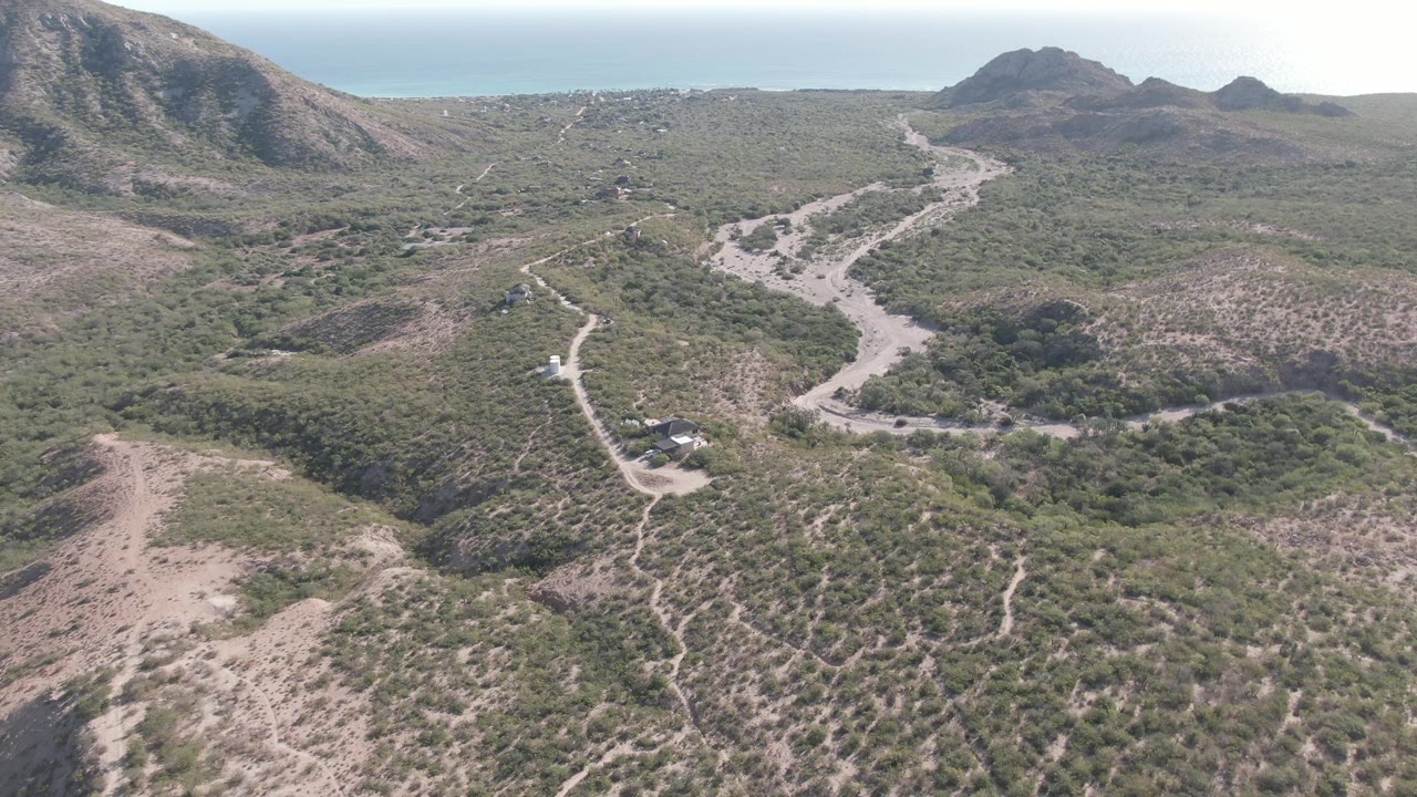 Opening aerial frame over Cabo Pulmo, Baja California Sur, showing a pale dirt road crossing scrubby hills toward distant mountains and ocean, with no visible people.