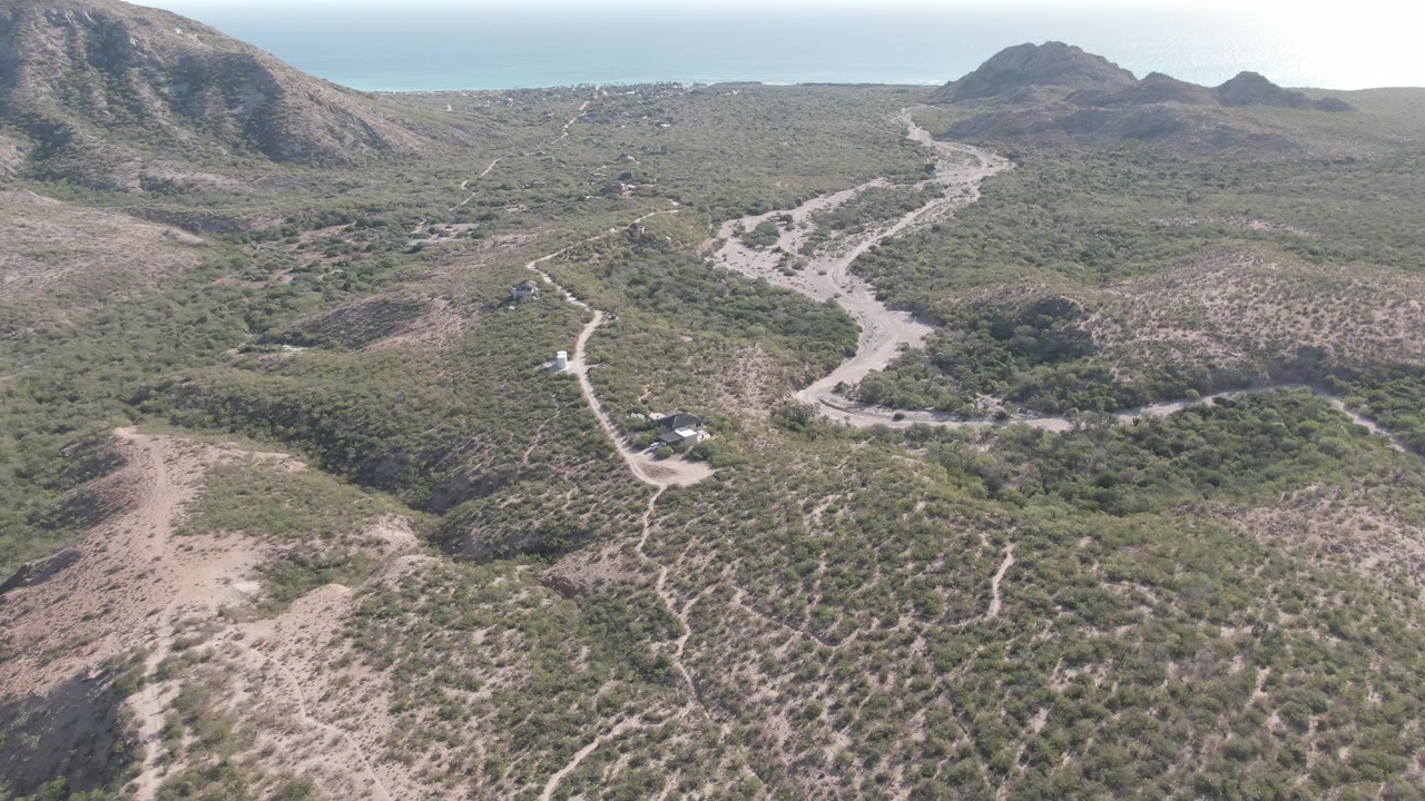 Aerial frame in Cabo Pulmo, Baja California Sur, with the dirt road and small vehicle slightly farther along the scrub-lined route below distant hills, no visible people.
