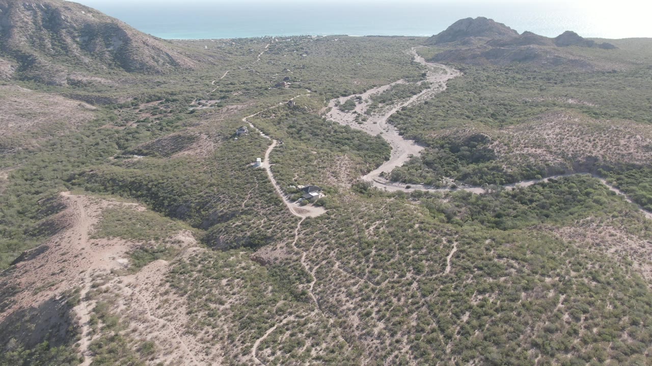 Wide Cabo Pulmo aerial view showing the curving dirt road threading through low green-brown vegetation, with mountains behind and the ocean horizon visible, no visible people.