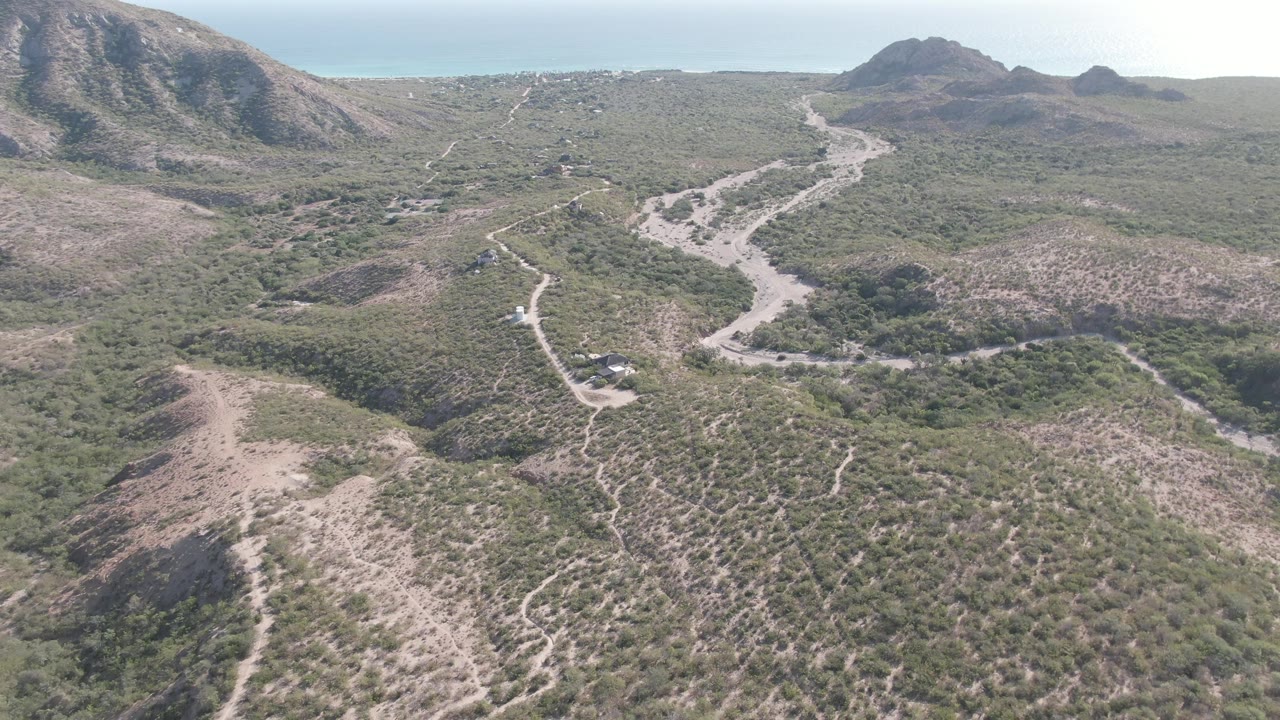 Aerial frame over Cabo Pulmo desert terrain, with a pale dirt road winding between scrub-covered slopes, rocky hills in the distance, and no visible people.