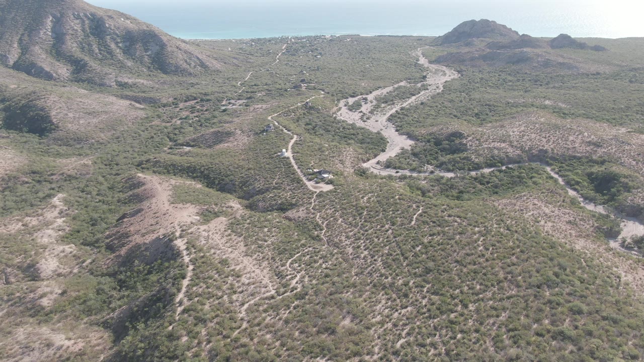 Drone view in Cabo Pulmo with the dirt road bending through green-brown scrubland, a vehicle near the road, rocky slopes, and no visible people.