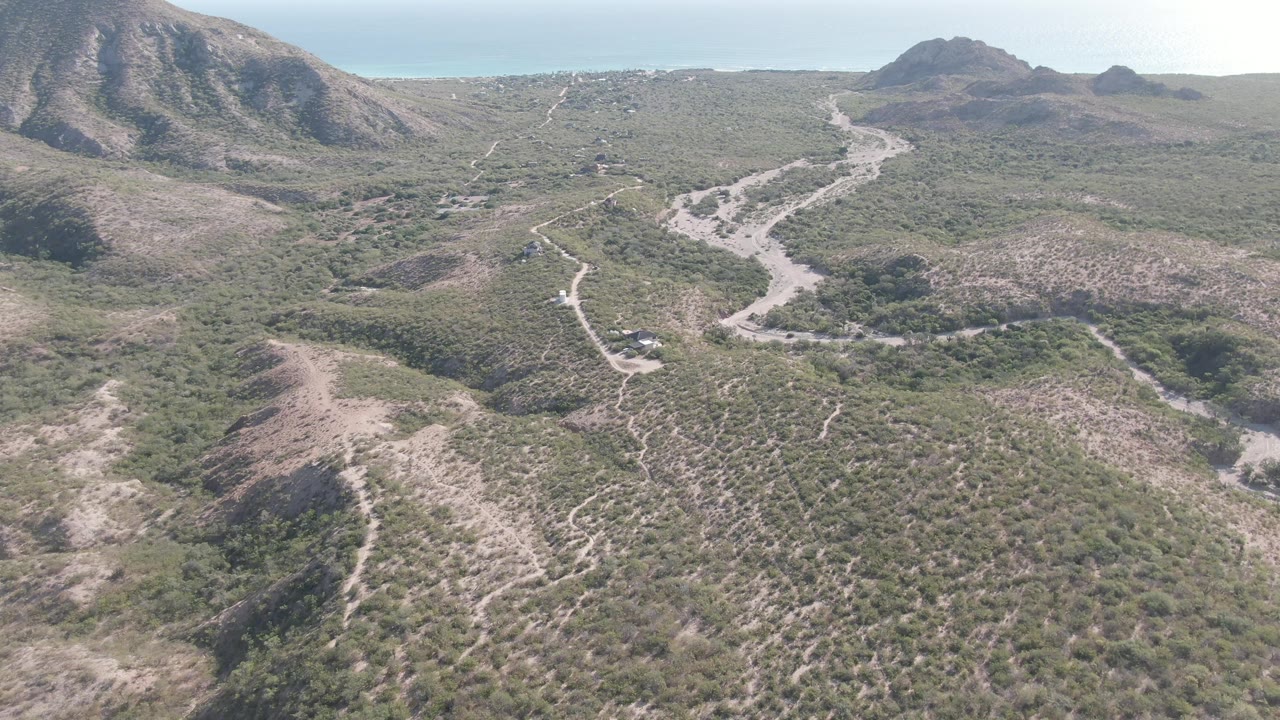 Aerial Cabo Pulmo frame with a vehicle on the central dirt road, low scrub in the foreground, hills on both sides, and no visible people.