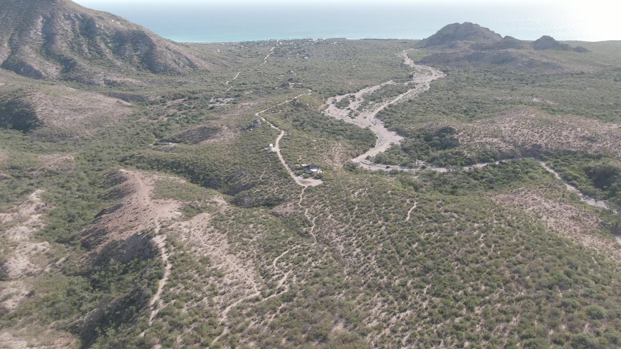 High aerial frame in Cabo Pulmo, Baja California Sur, with the road bending through tan and green brushland between rocky slopes and a broad mountain backdrop, with no visible people.