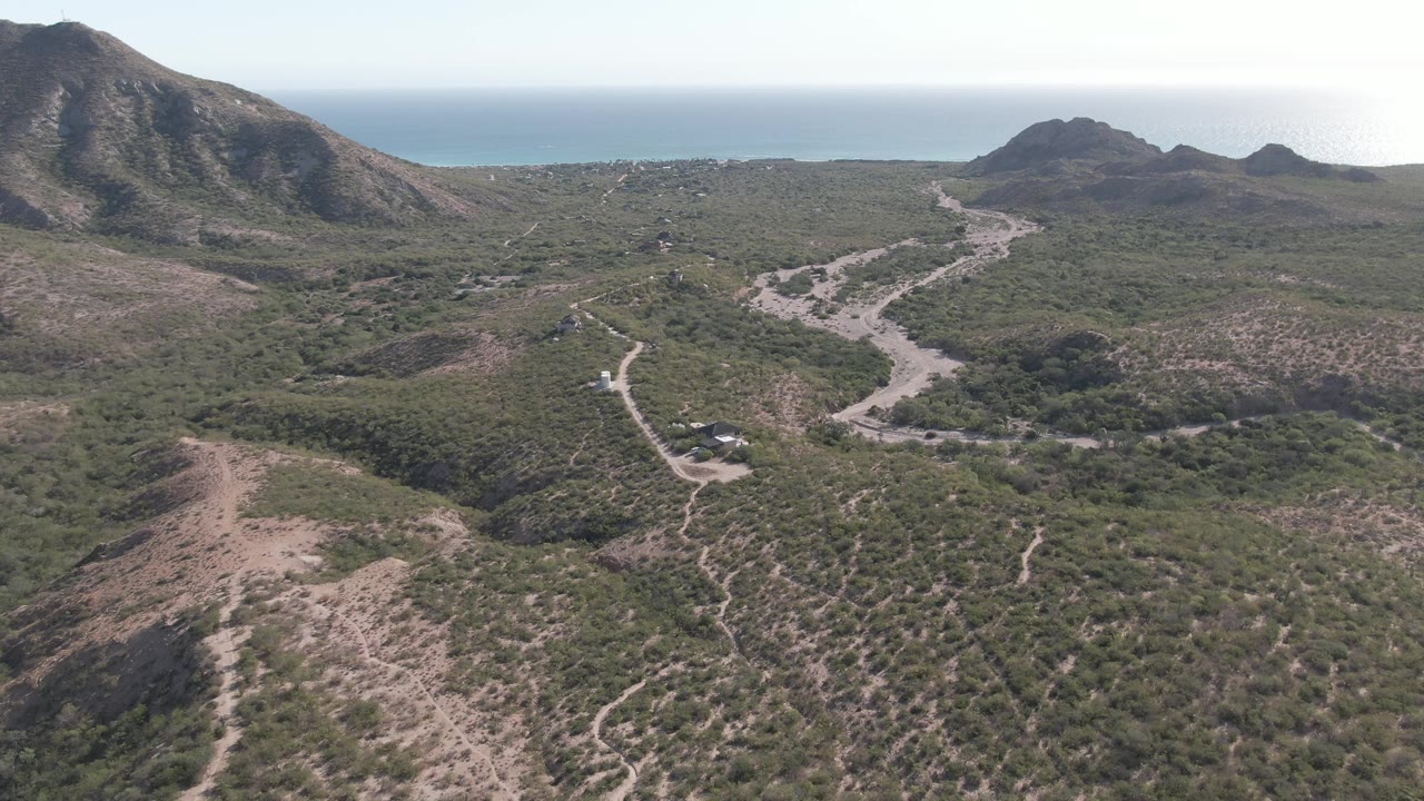 At two seconds, the aerial view holds on the winding Cabo Pulmo road, with a rocky slope on the left, rounded hills on the right, the blue sea behind them, and no visible people.