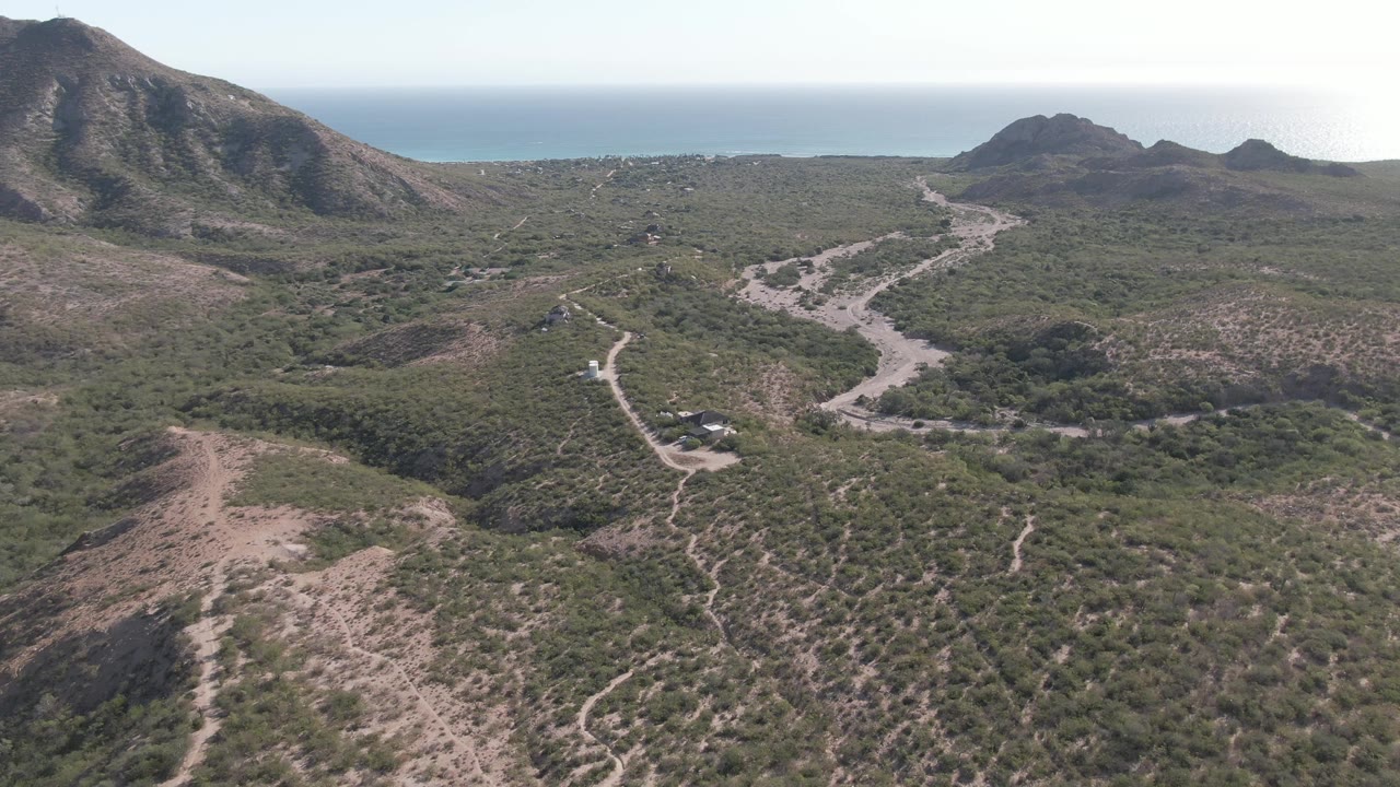 Three seconds in, the Cabo Pulmo road remains visible below the drone, with a low foreground ridge, dense patches of green brush, distant hills, ocean horizon, and no visible people.