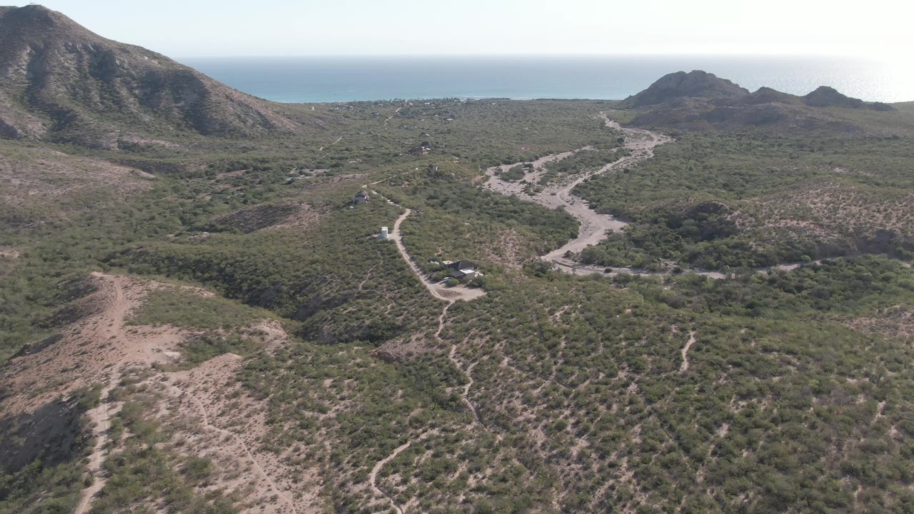 Aerial view of a winding dirt road through dry hillside scrub with ocean beyond.