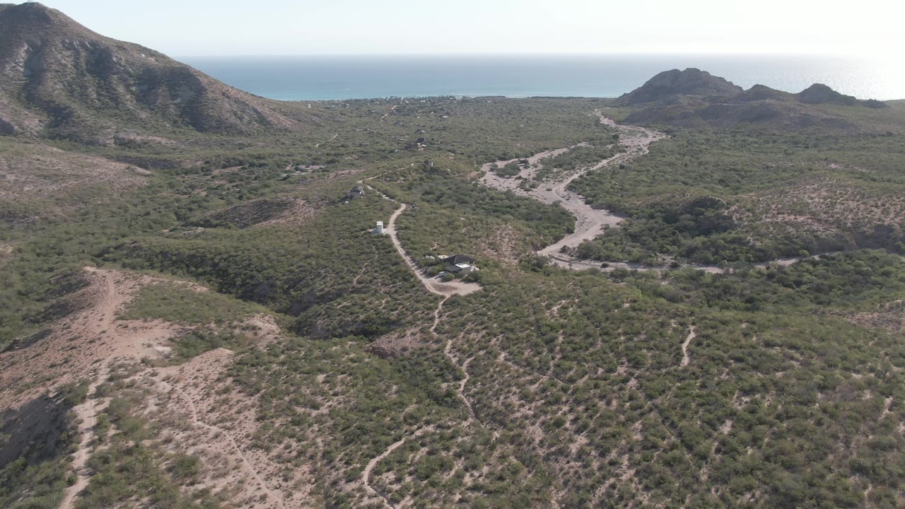 Aerial frame in Cabo Pulmo with the vehicle visible on the lower road, scrubby hills across the foreground, dark mountain slopes to the sides, distant ocean, and no visible people.