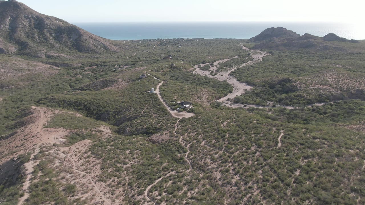 High aerial view in Cabo Pulmo, Baja California Sur, with branching dirt tracks crossing dry scrub, rocky hills in the distance, ocean beyond them, and no visible people.