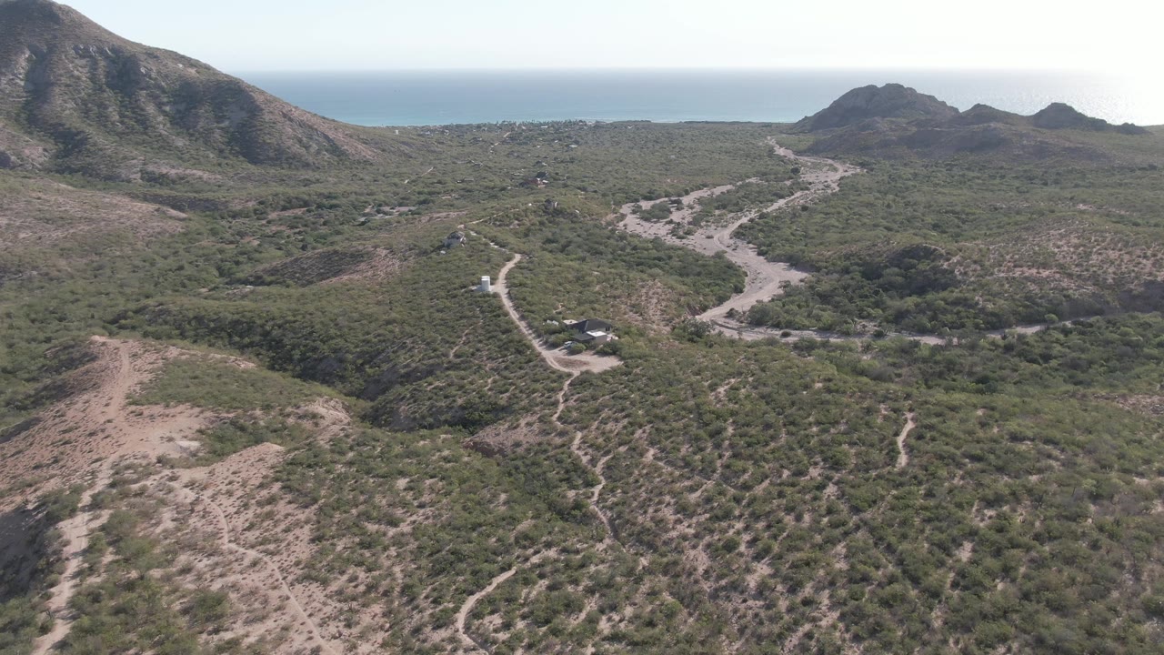 Cabo Pulmo aerial frame with a pale dirt road curving through low vegetation, a small vehicle near the lower center-left area, mountain slopes, distant sea, and no visible people.