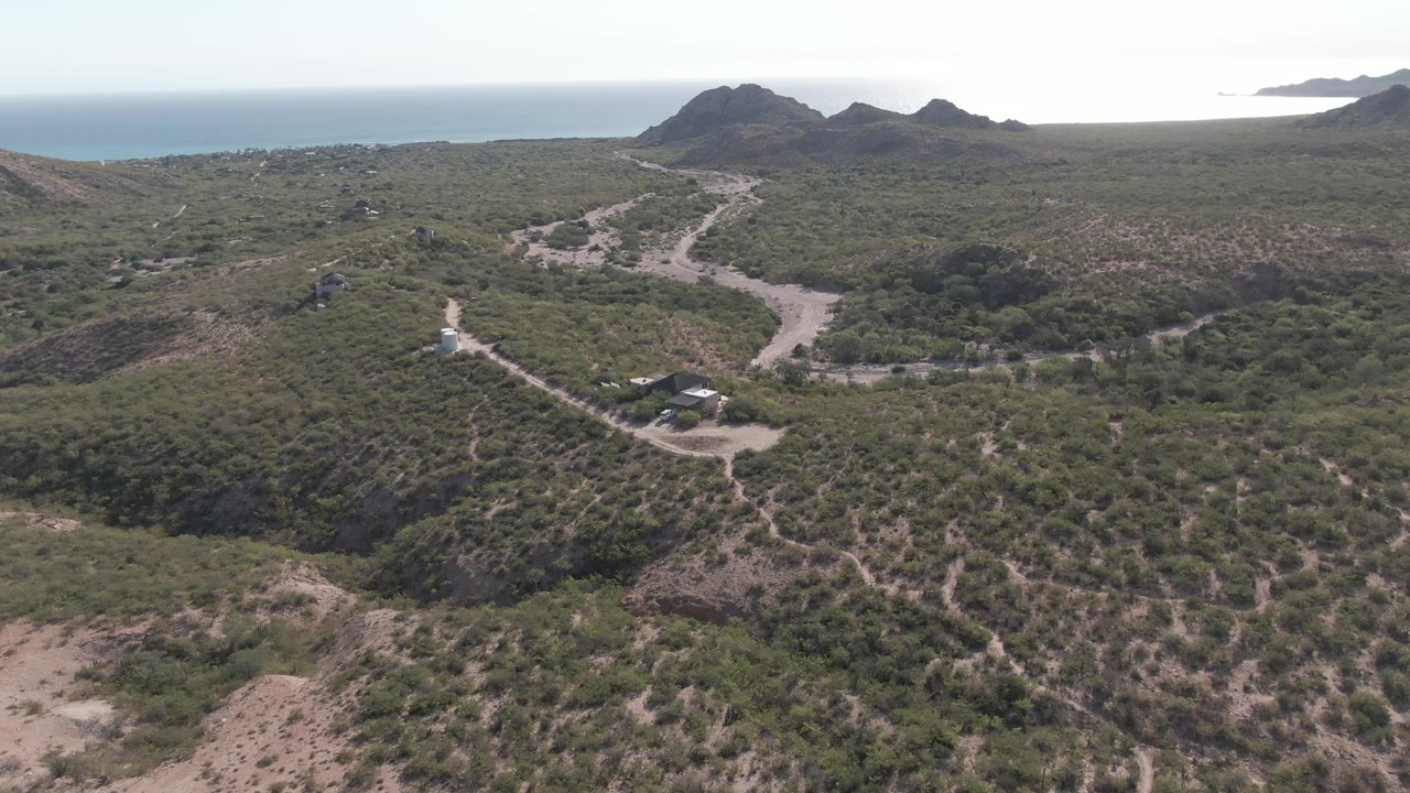 Aerial view in Cabo Pulmo, Baja California Sur, Mexico showing a white vehicle on a pale dirt road through scrub-covered hills with the ocean on the horizon and no visible people.