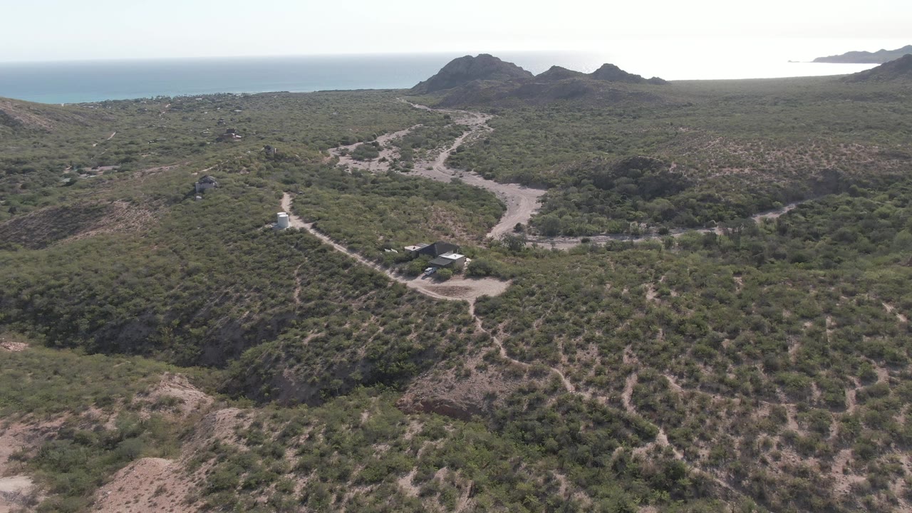 Wide aerial frame in Cabo Pulmo, Baja California Sur, Mexico with the white vehicle farther along the dirt track near a branching road, surrounded by scrub hills and distant ocean, with no visible people.