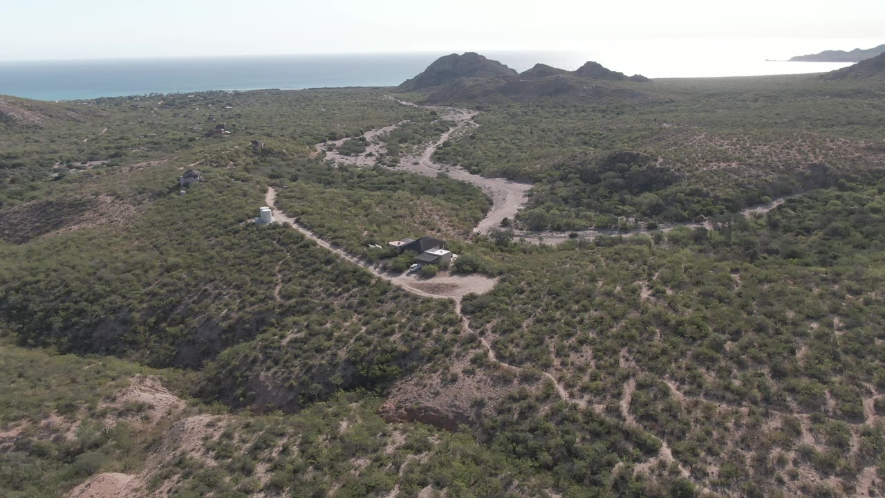 Aerial frame in Cabo Pulmo, Baja California Sur, Mexico showing the white vehicle continuing along the road below low dark hills, with green-brown vegetation, ocean, and no visible people.