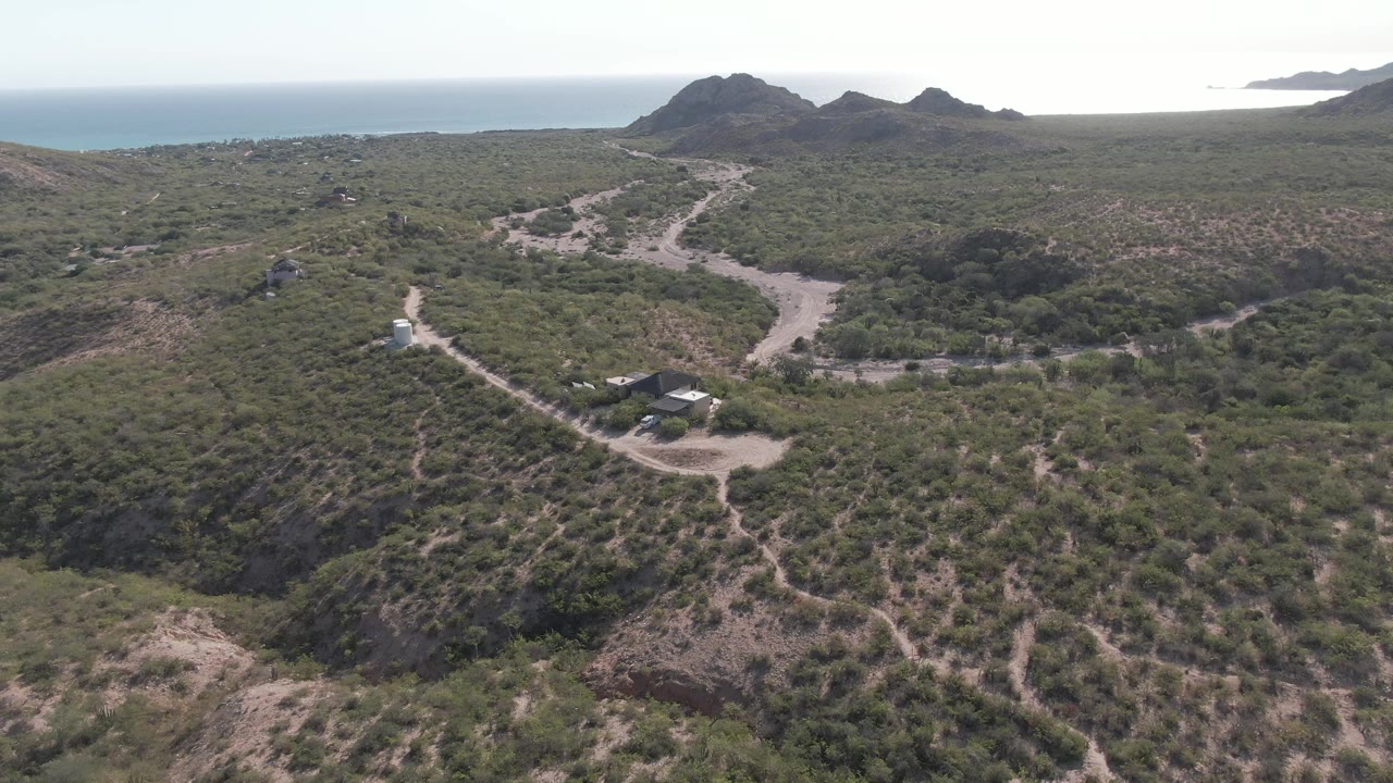 High aerial view of a winding dirt road, scrubby hills, and a white vehicle near small structures.