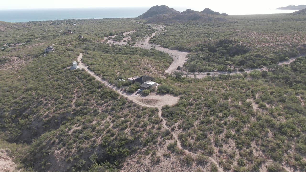 Drone view over Cabo Pulmo hillside scrub with a winding dirt road, small structures below, distant hills and sea on the horizon, and no visible people.