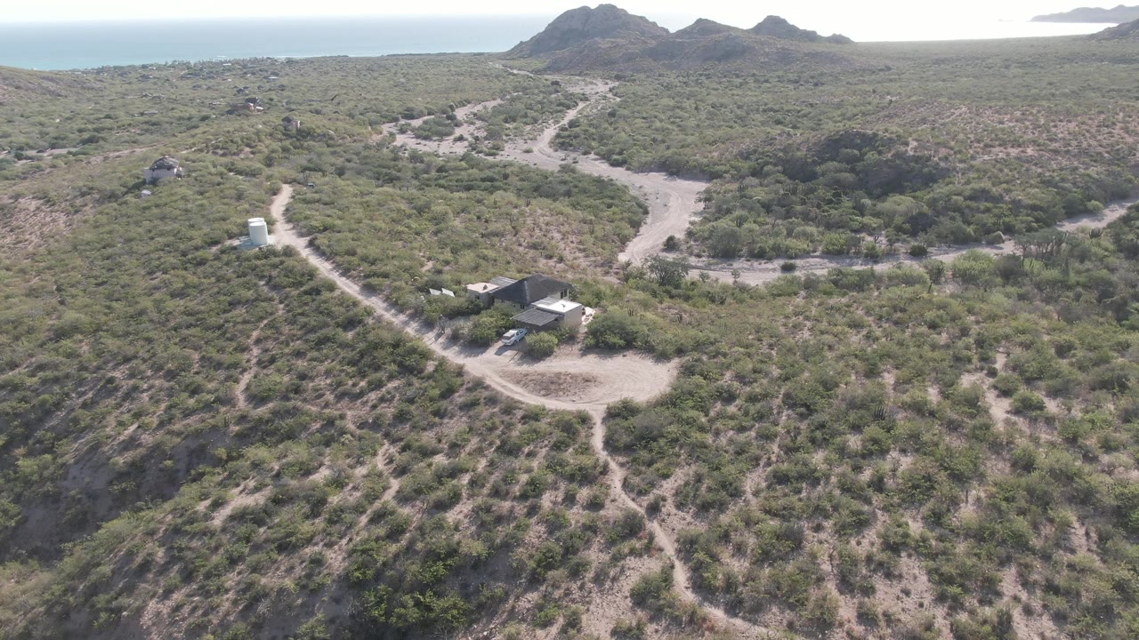Aerial frame from Cabo Pulmo showing the road curving around scrubland, a small building cluster near the lower edge, distant rocky hills, sea, and no visible people.