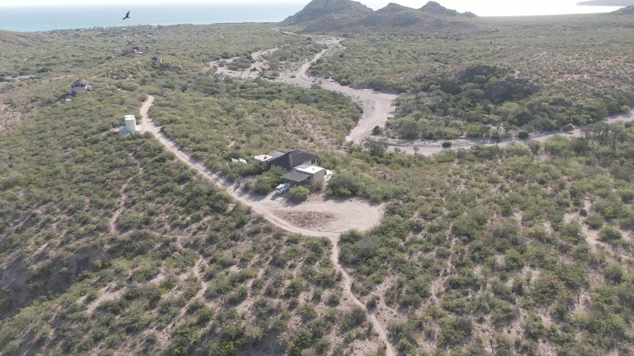 Cabo Pulmo aerial view with a pale dirt track, sparse green-brown vegetation, a small trailer or structure area below, distant water, and no visible people.