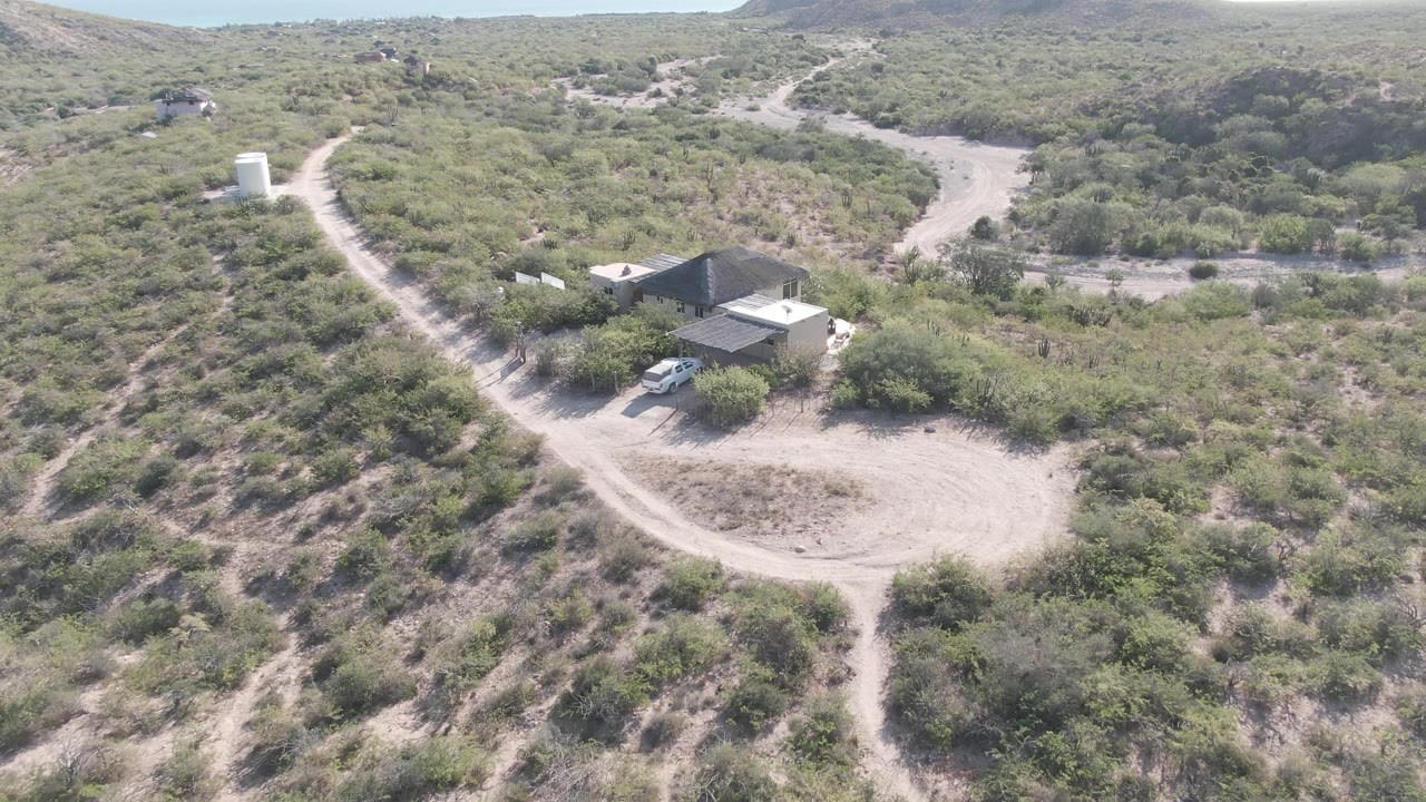 Elevated Cabo Pulmo frame with intersecting dirt roads, a low desert house, a white vehicle by the structure, green shrubs, and no visible people.