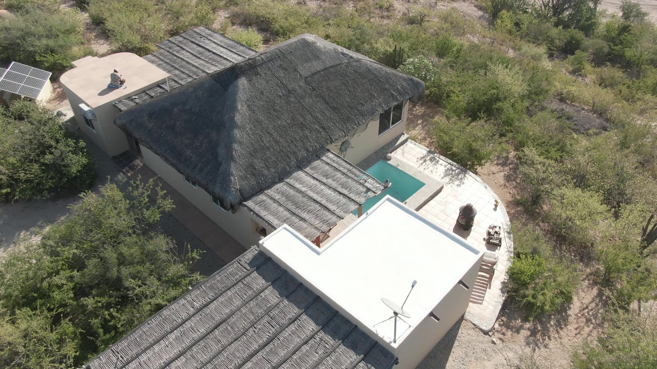 Aerial frame over a Cabo Pulmo desert house with a thatched roof, adjacent solar panels, scrub vegetation, pale dry ground, and no visible people.