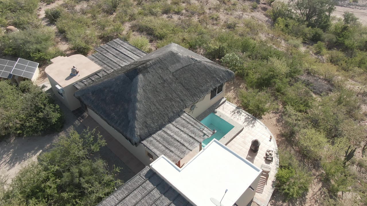 Drone view over a thatched-roof house, pool, solar panels, and desert brush.
